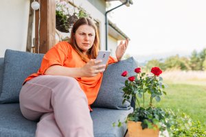 Young redhead woman relaxing with phone in the yard of the house in summer. Negative people emotion. Upset 30s women is using a smartphone